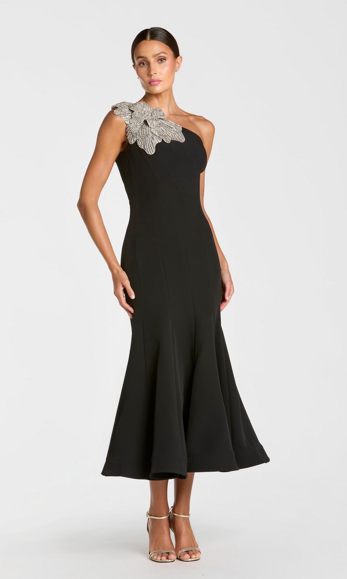 A woman stands in neutral heels against a plain light background, dressed in the Mac Duggal 13221 Tea-Length Party Dress in black, which features one-shoulder styling and a silver embellished floral applique at the shoulder.
